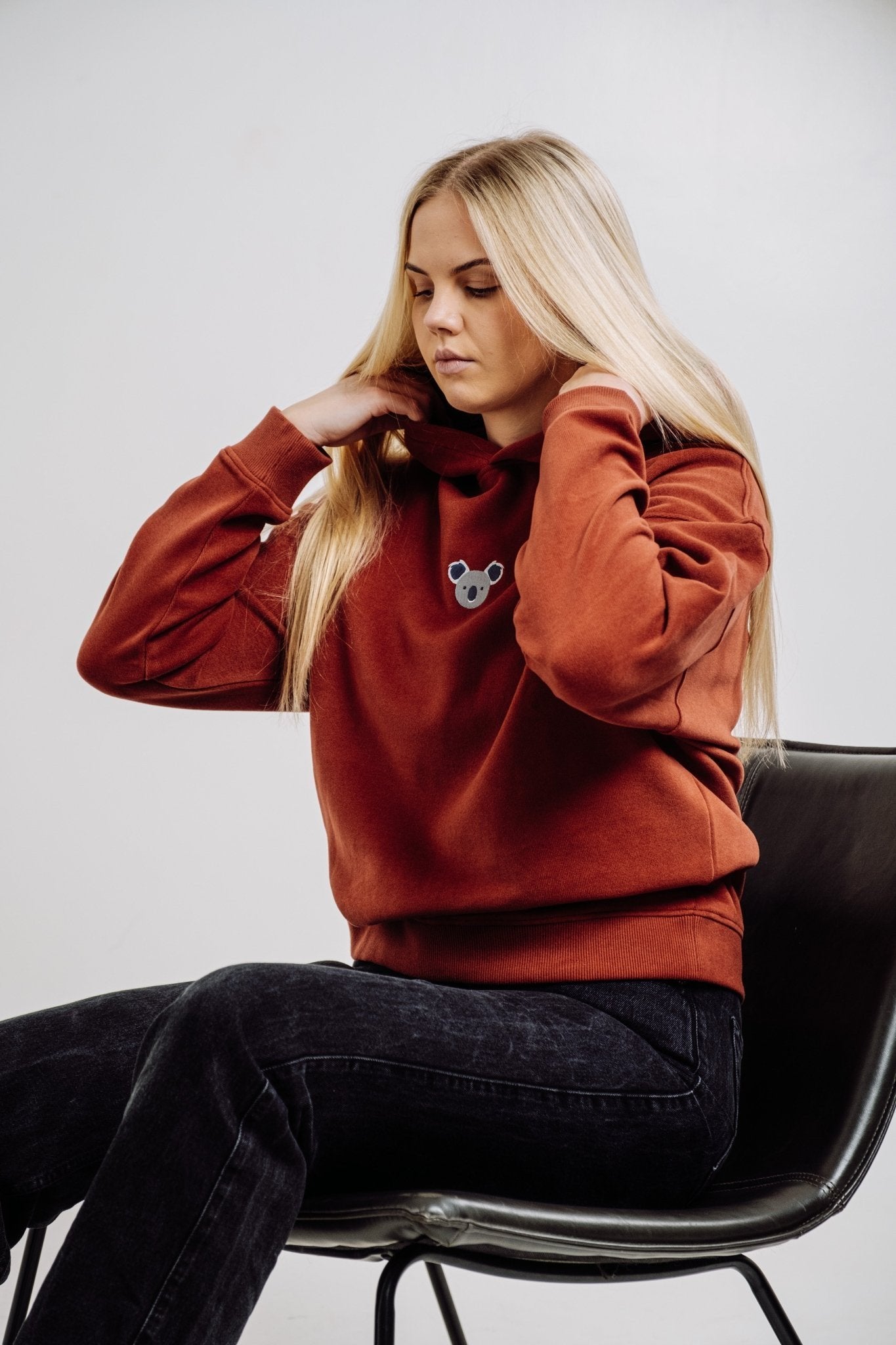5. Woman adjusting hood of Sossu Koala Hoodie in terracotta, seated in chair, studio setting
