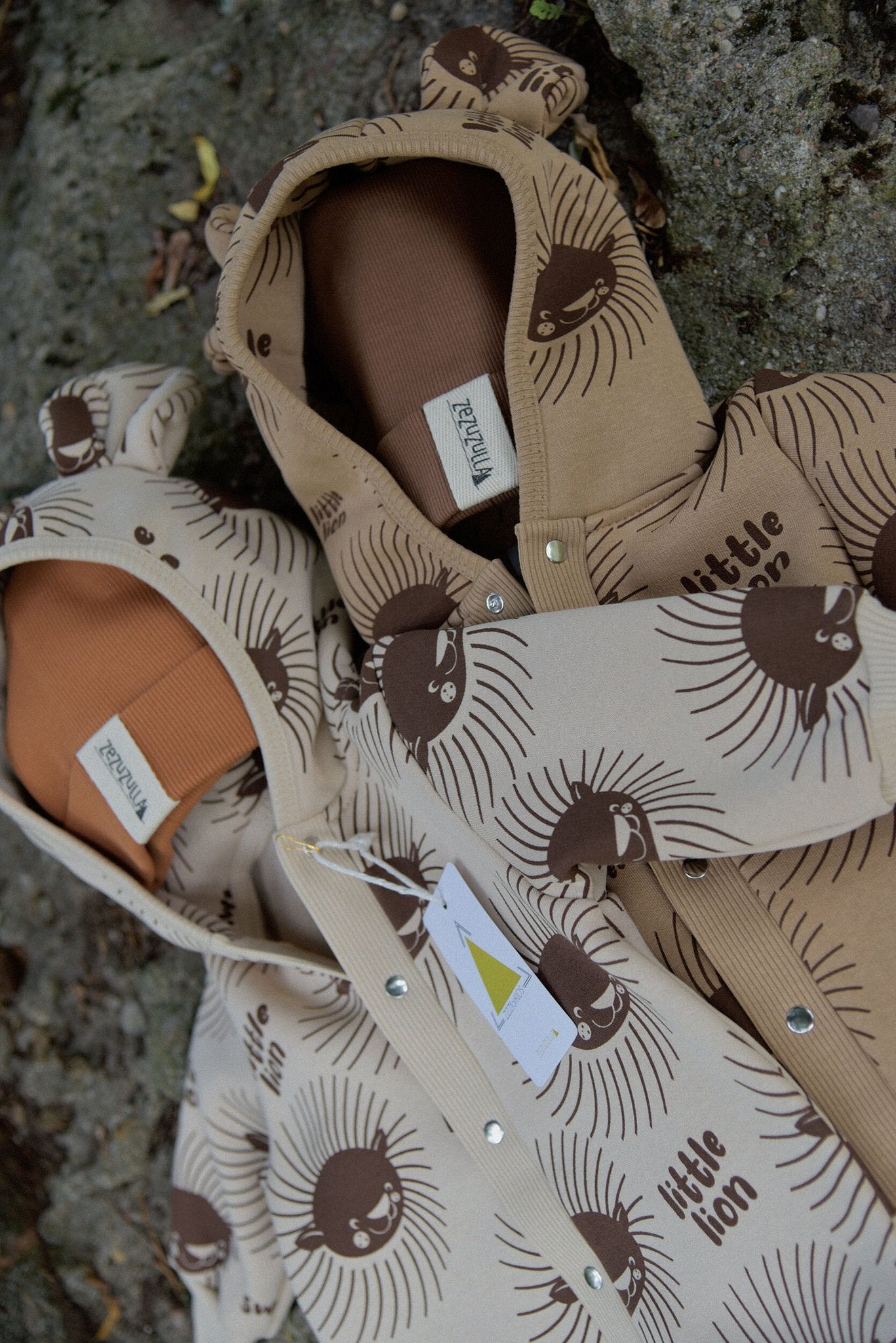 1. Two baby jumpsuits with lion print and hood with ears, in beige and brown, displayed outdoors on a rock