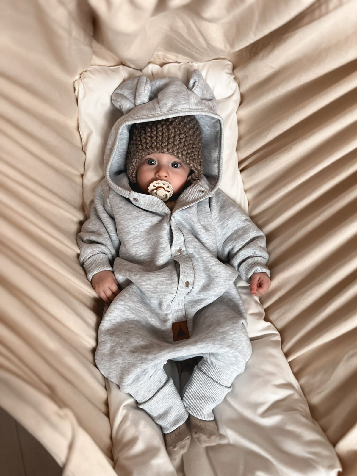 1. Baby wearing Zezuzulla melange grey eared jumpsuit with hood lying on cream fabric