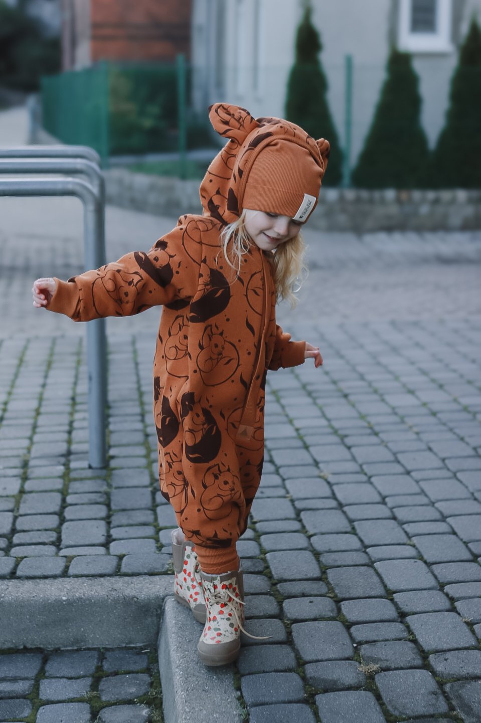 1. Child balancing on curb in cinnamon squirrel jumpsuit with hood and ears, outdoors