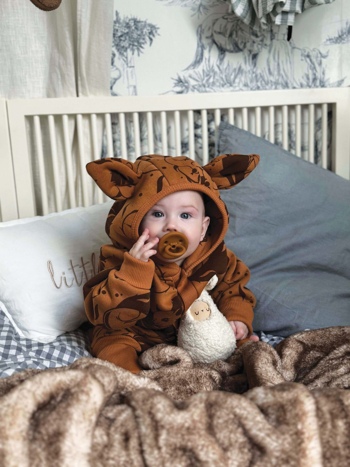 1. Baby sitting in crib wearing cinnamon squirrel jumpsuit with hood and ears, holding toy