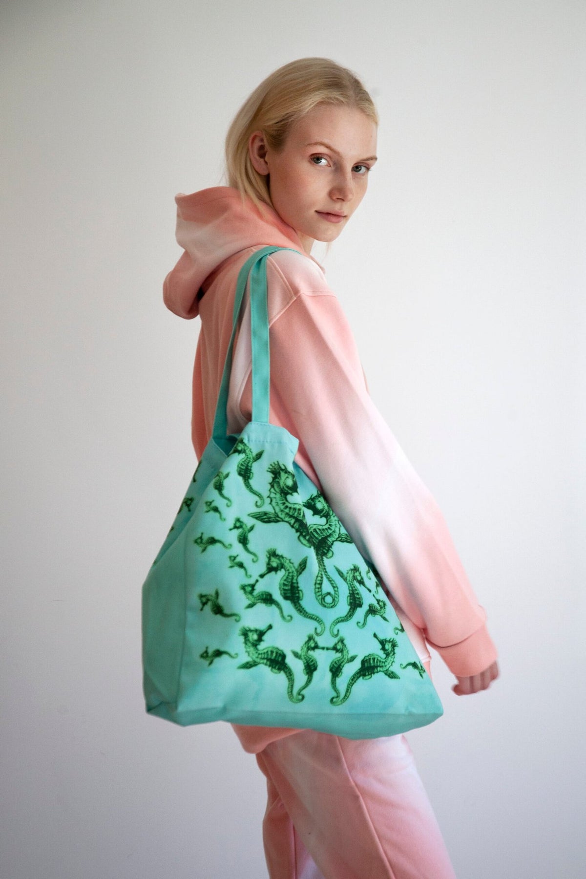 2. Woman in pink and white hoodie carrying Tanel Veenre Mirage tote bag featuring green seahorse pattern