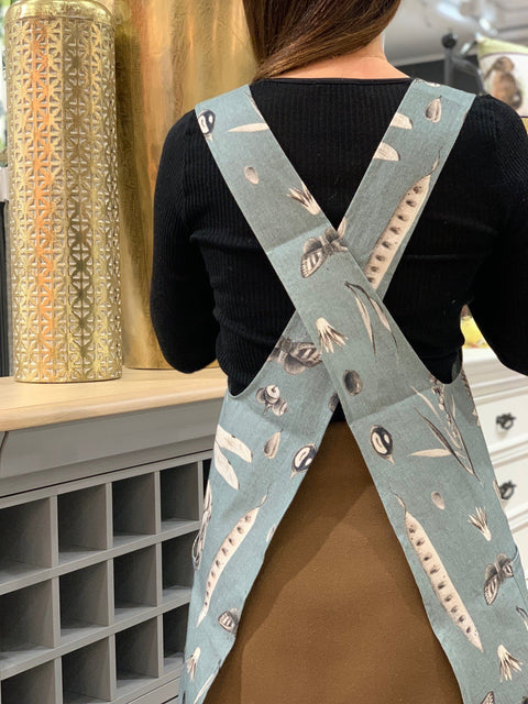 1. Woman wearing Seeds Aqua print apron with crossed straps, shown from back in a home setting