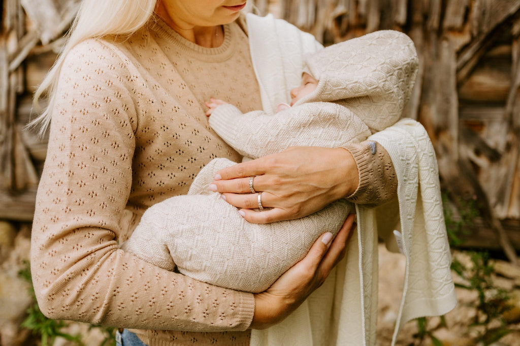 1. Woman holding baby wrapped in beige merino wool blanket and romper with matching bonnet outdoors