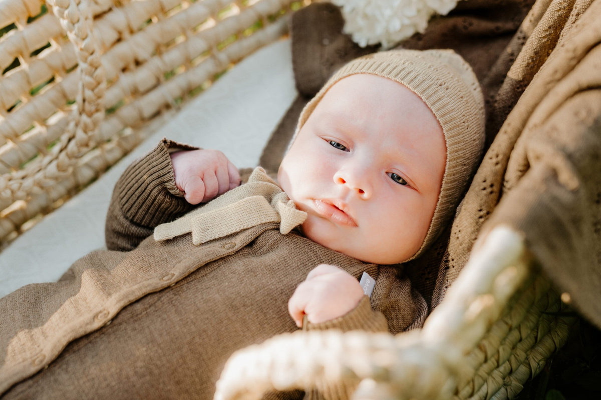 4. Baby in brown merino wool romper and bonnet lying in a basket with a braided blanket