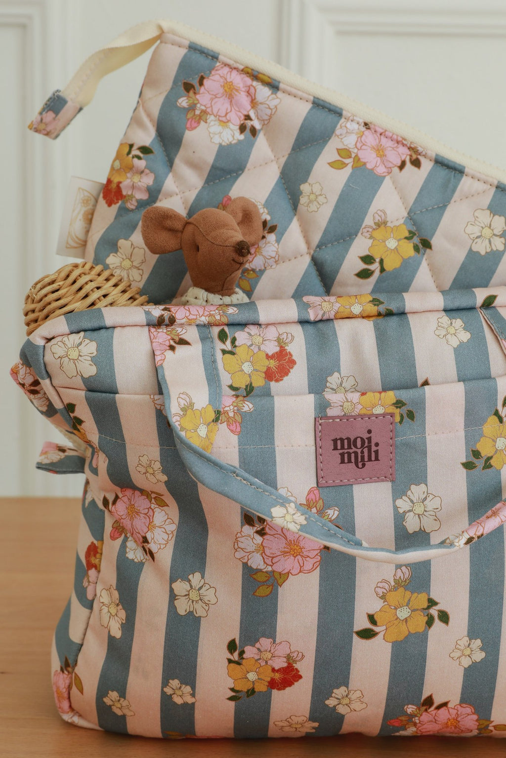 2. Close-up of blue striped makeup bag with floral design and Moi Mili logo, small toy peeking out, on wooden surface