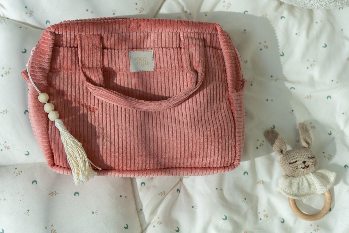 1. Large coral makeup bag with handles and tassel detail, on a patterned cushion with a toy