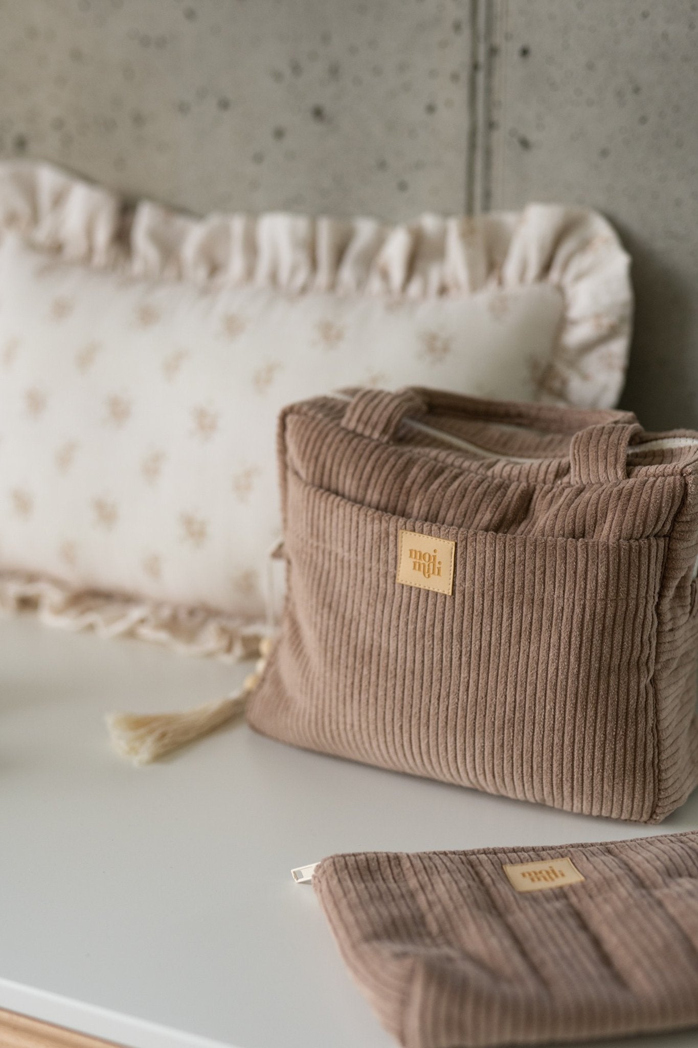 1. Latte-colored large makeup bag with Moi Mili logo on a white surface, next to a decorative pillow, featuring tassel detail.