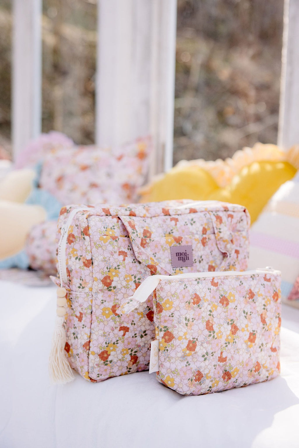 1. Moi Mili large and small floral makeup bags on bed with colorful pillows in background
