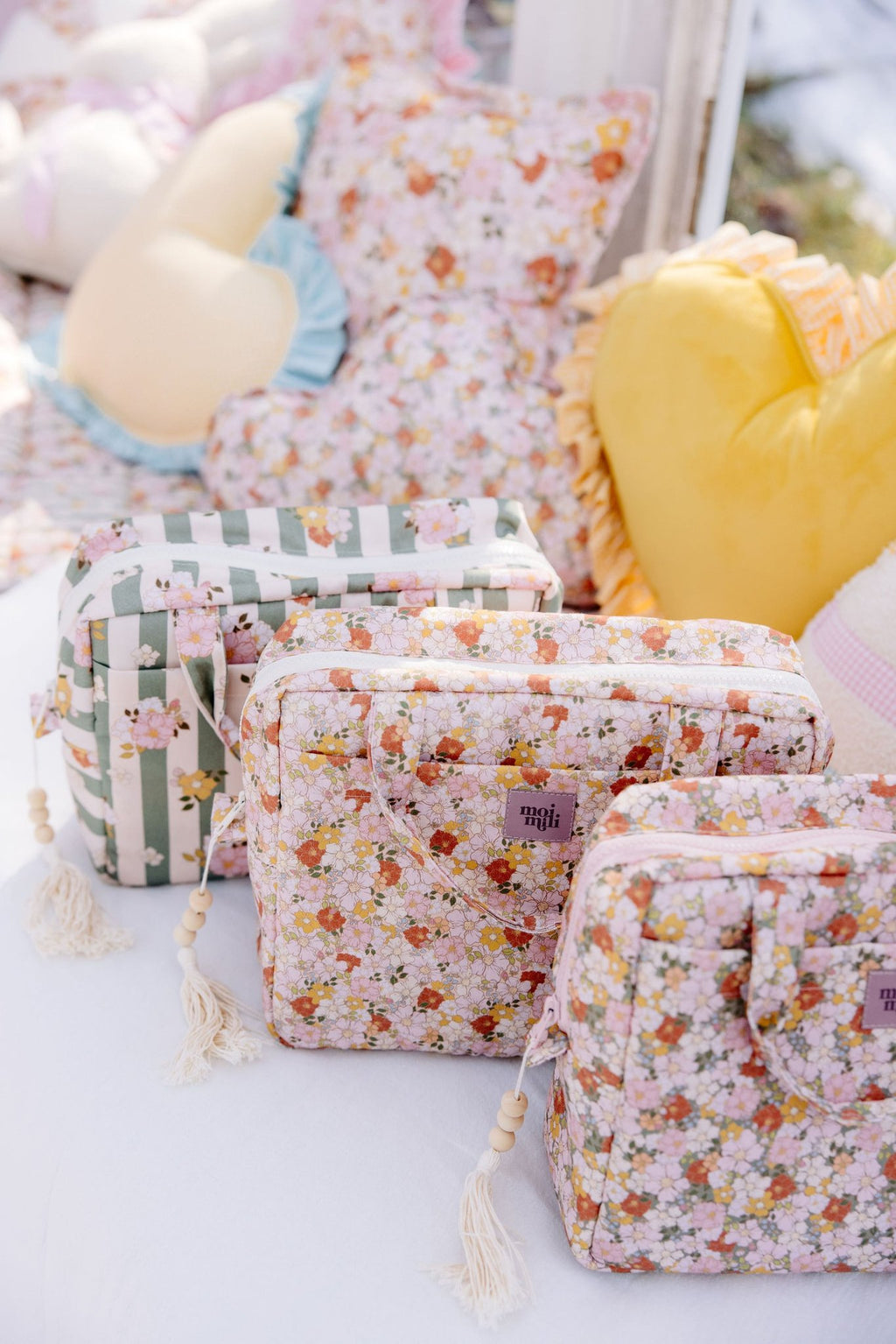 1. Three Moi Mili floral and striped makeup bags on bed with decorative pillows in background