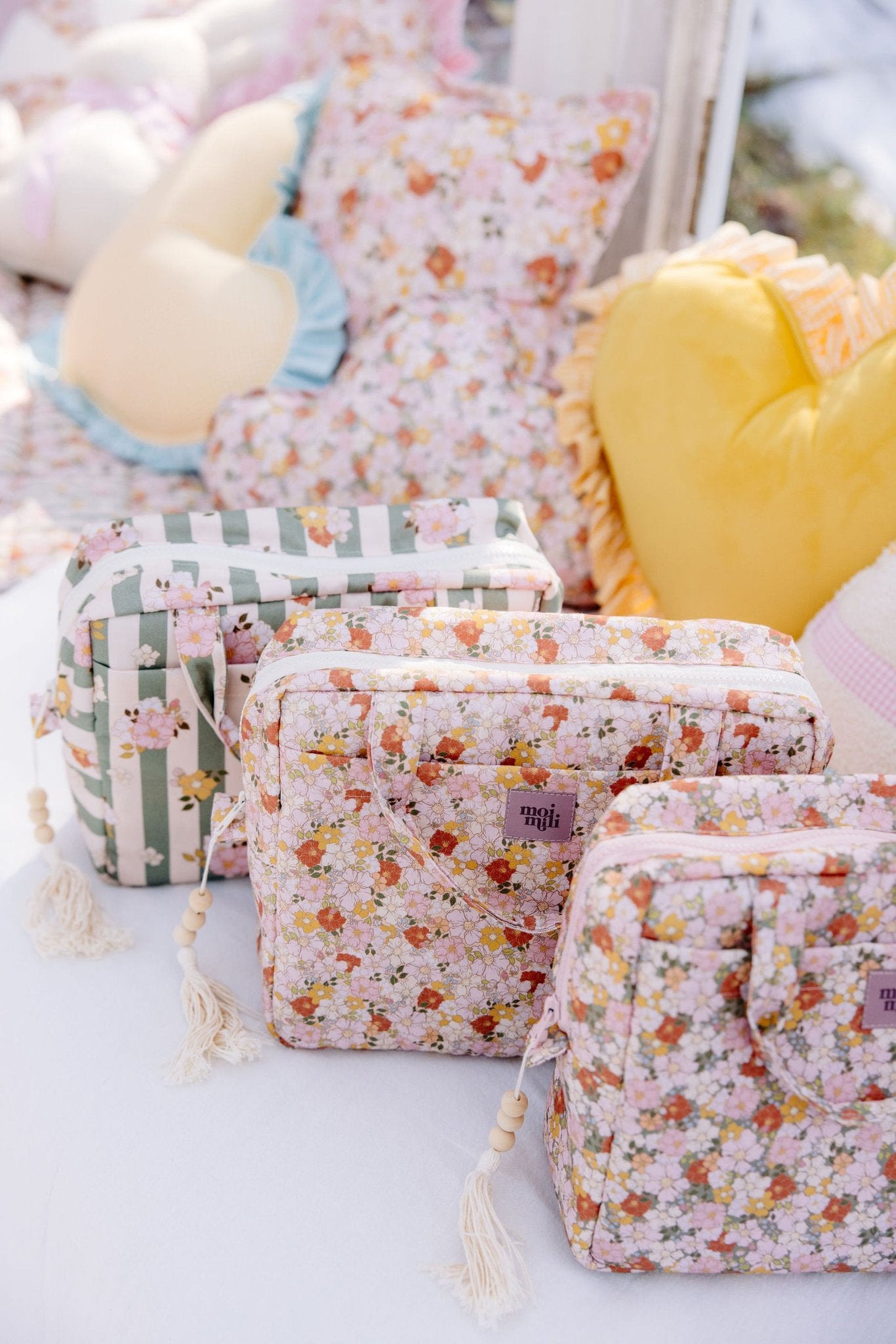 1. Three Moi Mili floral and striped makeup bags on bed with decorative pillows in background