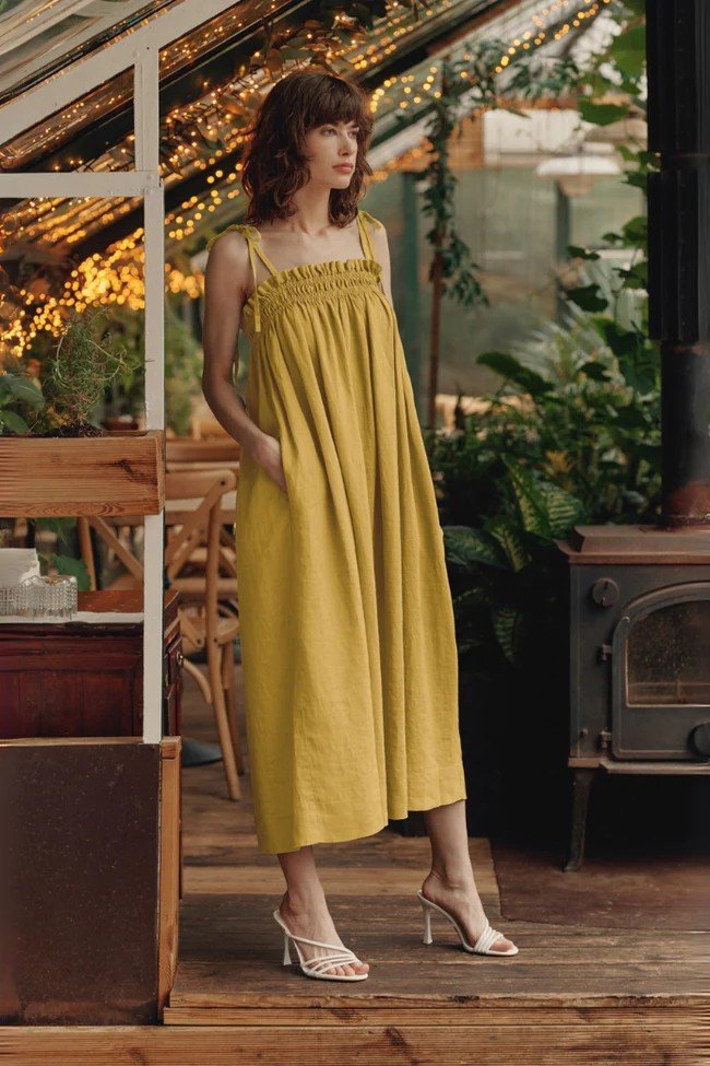 1. Woman wearing a lemon curry linen dress with adjustable straps and inseam pockets, standing in a cozy indoor setting with plants and wooden furniture
