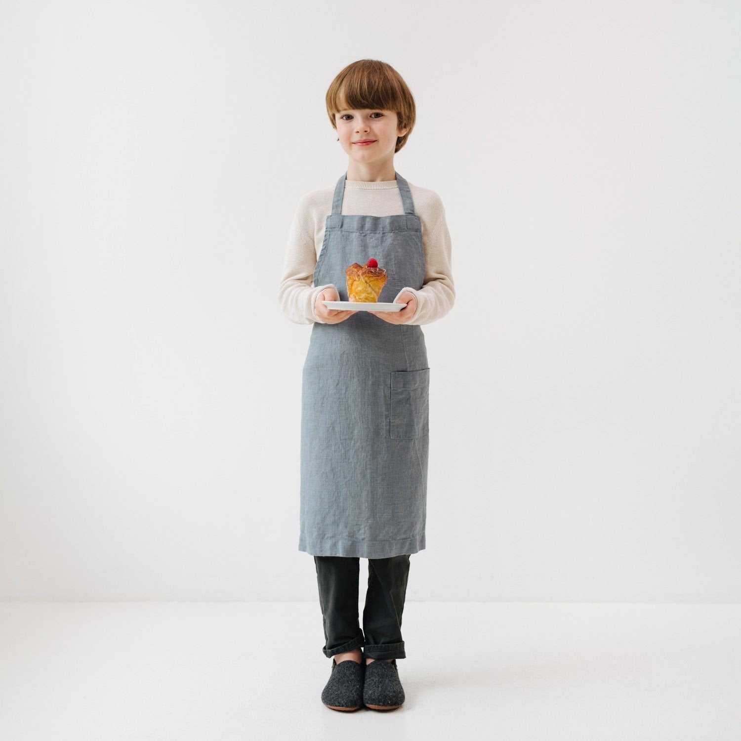 1. Child wearing blue fog linen apron holding a plate with dessert, standing in a bright room