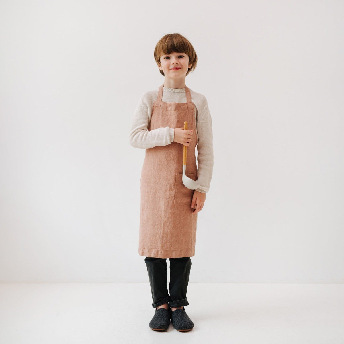 1. Child wearing cafe creme linen apron holding a ladle, standing against a white background