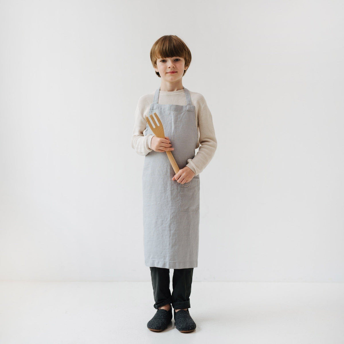 2. Child holding wooden kitchen tool while wearing light grey linen apron