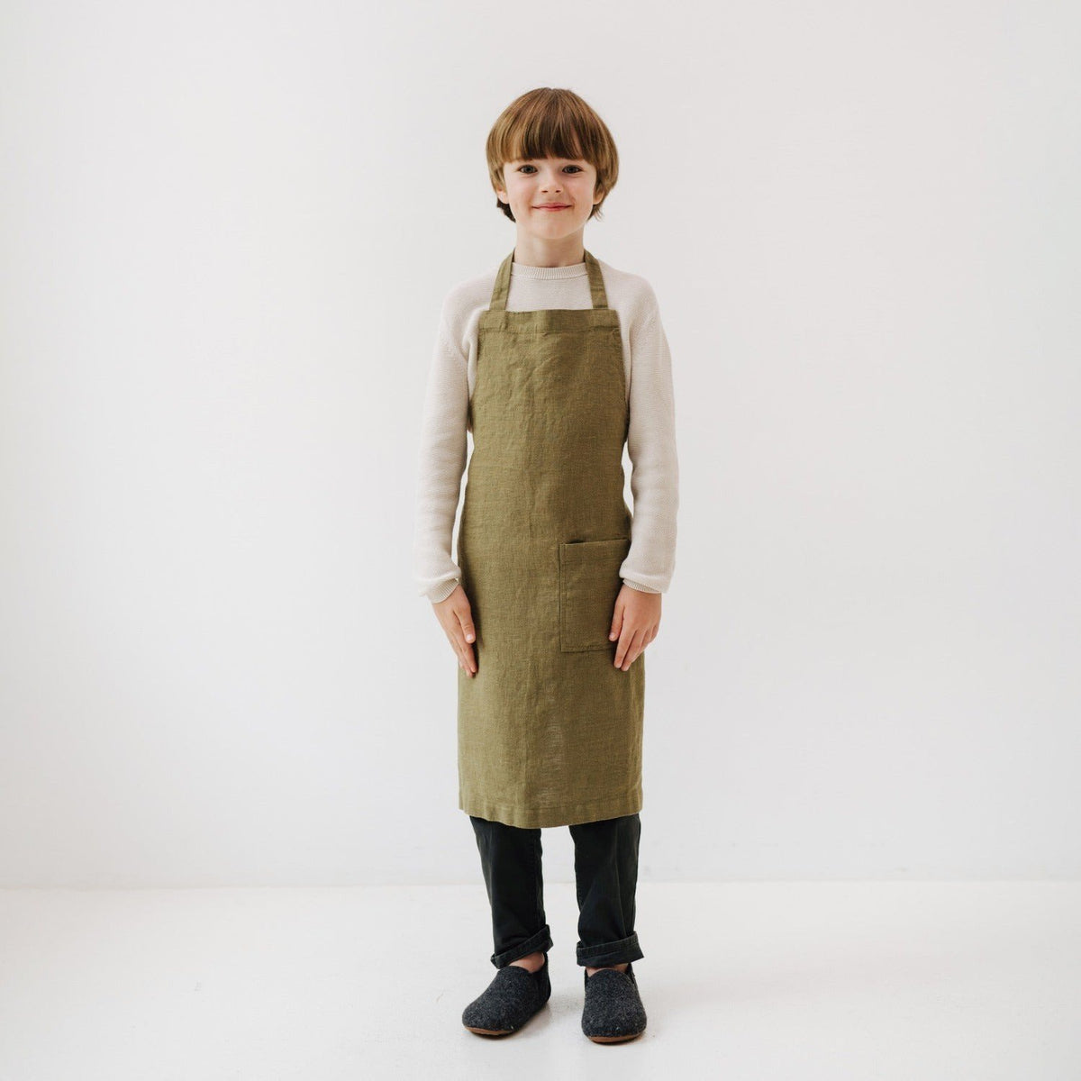 3. Child wearing olive green linen apron with front pocket, standing with hands at sides in a white room