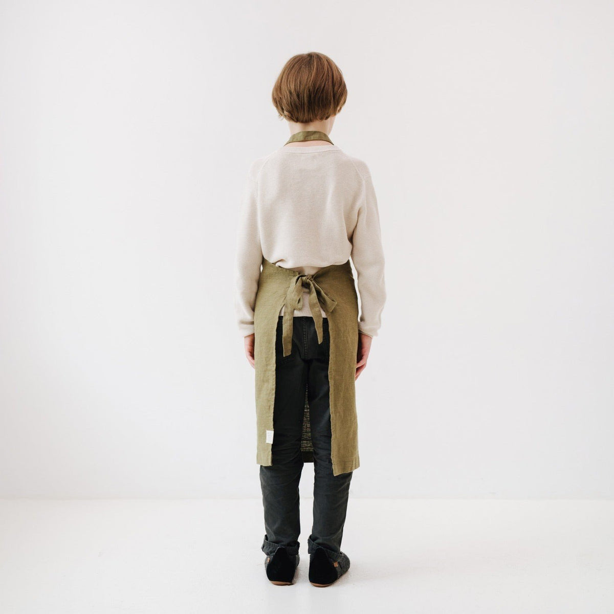 5. Back view of child wearing olive green linen apron with tied straps, standing in a white room