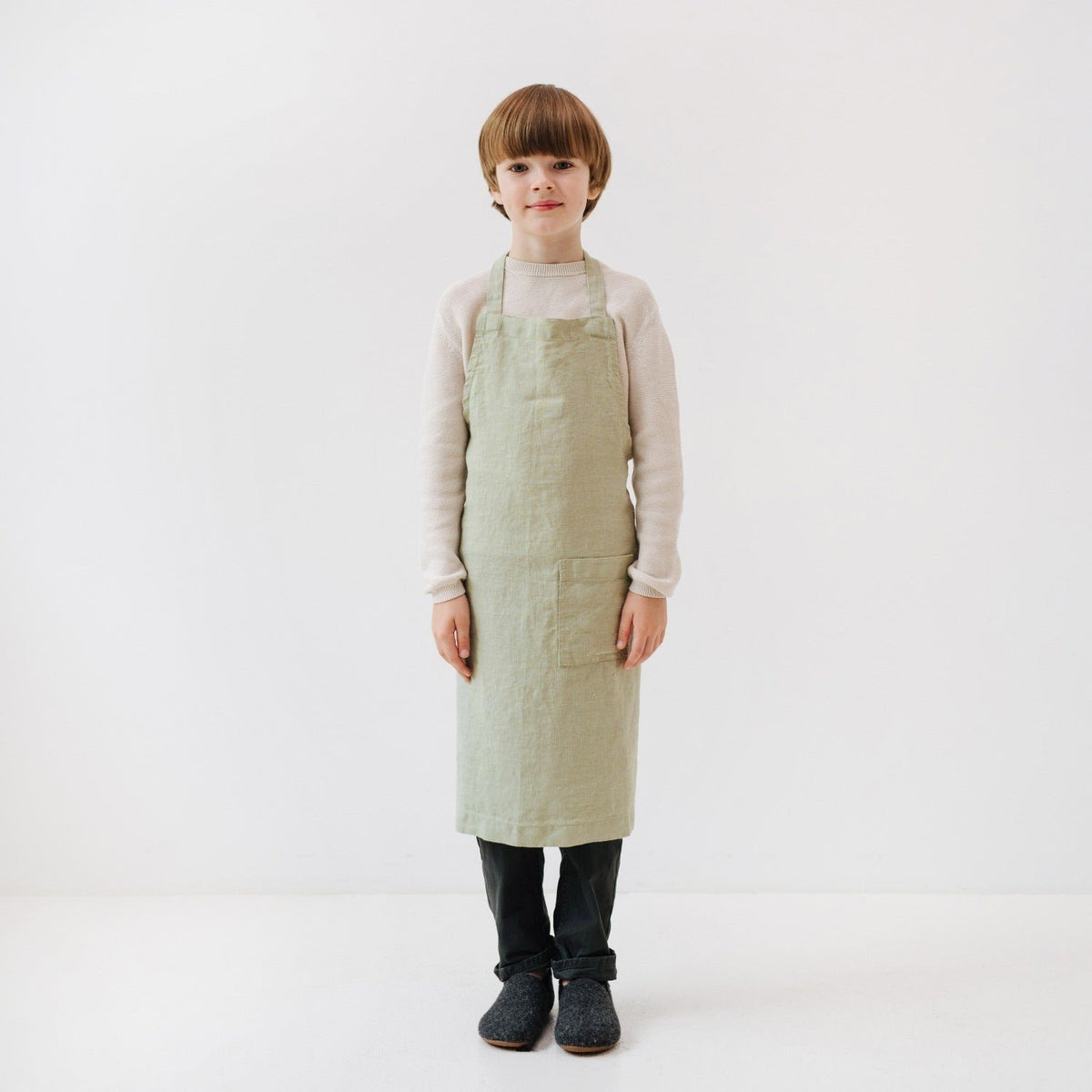3. Front view of child wearing sage green linen apron with front pocket, standing in white studio