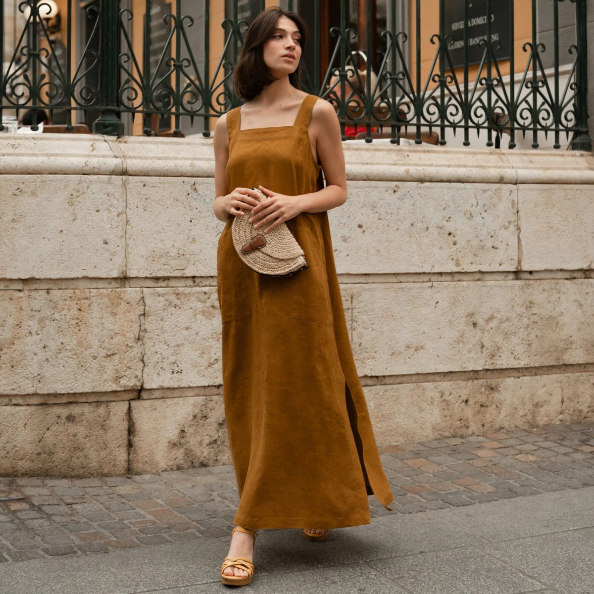1. Woman wearing bronze linen A-line dress with wide straps and square neck, holding a woven clutch, walking on a cobblestone street