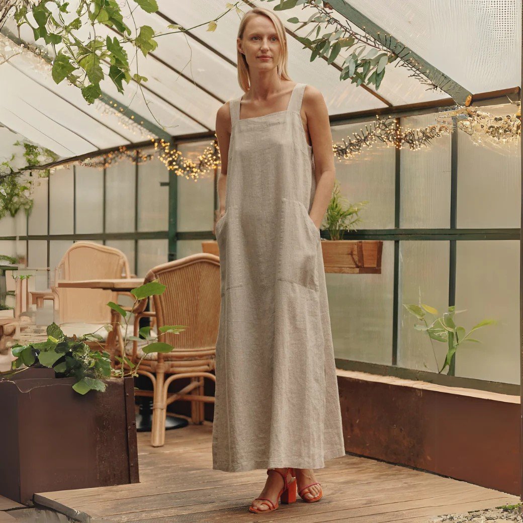 1. Woman wearing a long linen dress with wide straps and square neck, standing in a greenhouse setting with plants and wicker furniture