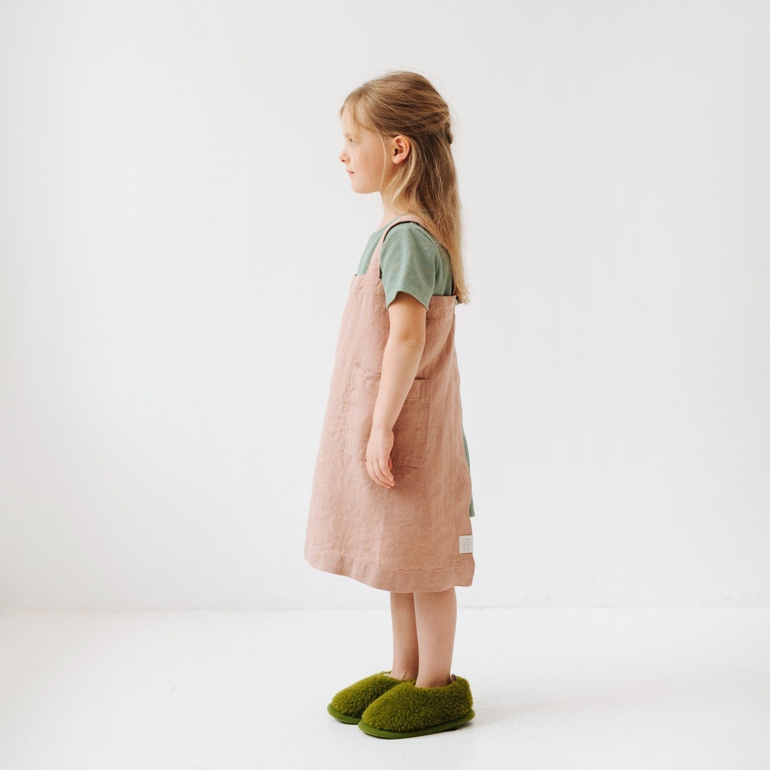 3. Side view of girl in pink linen pinafore apron with green shirt and green slippers in studio setting