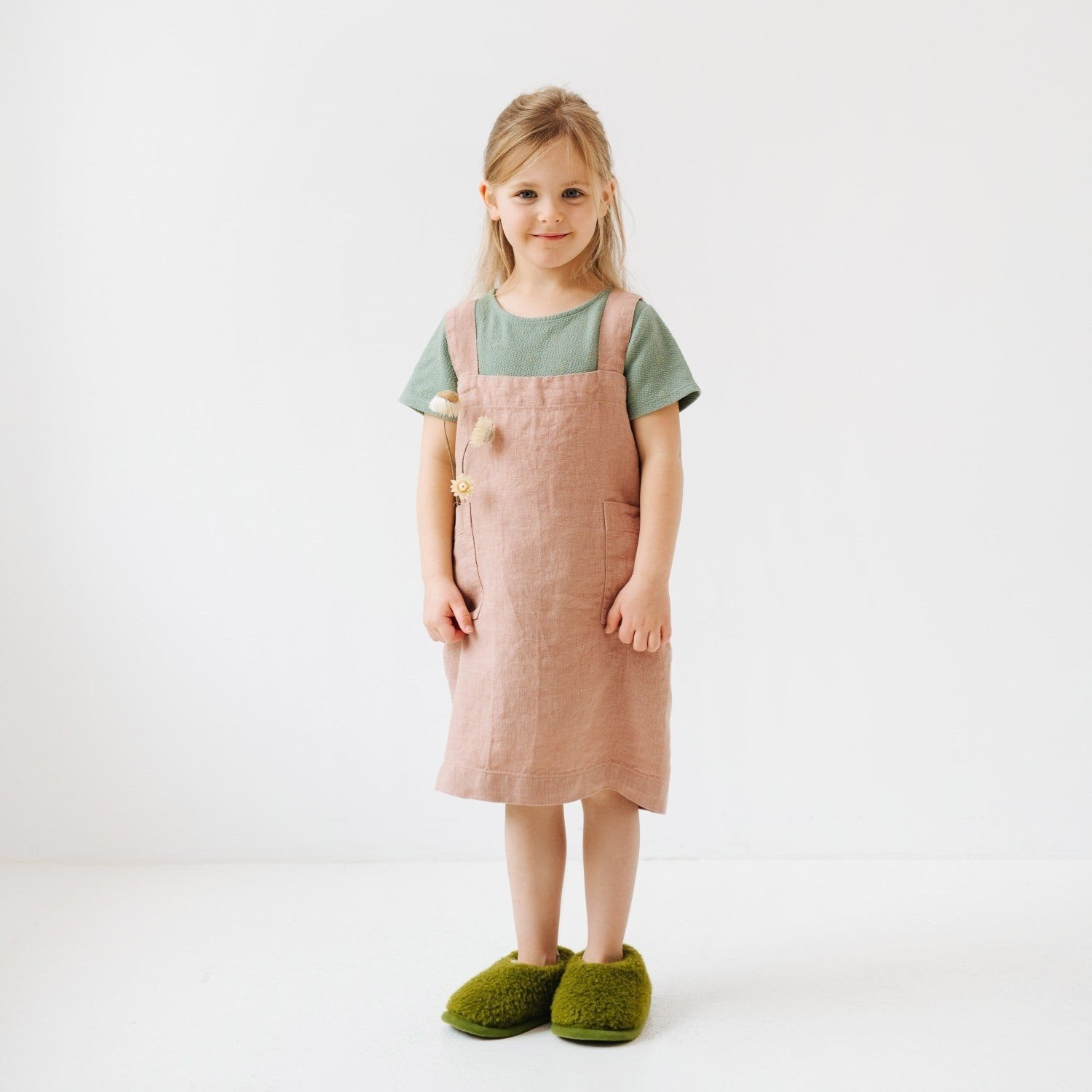 5. Girl in pink linen pinafore apron with green shirt and green slippers holding flowers in studio setting
