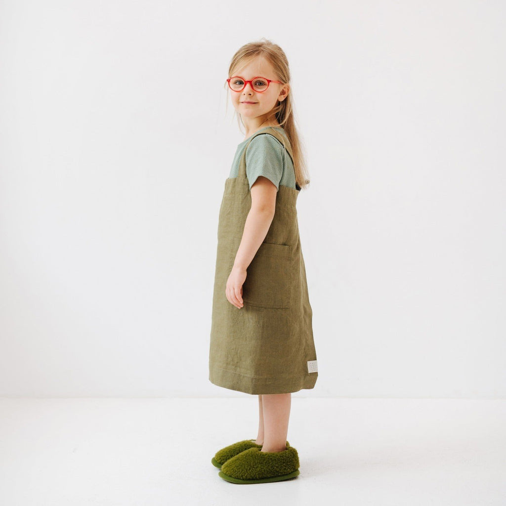 4. Side view of girl wearing olive linen pinafore apron, standing on a white background