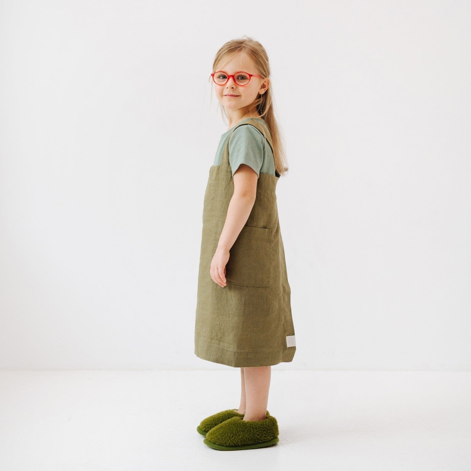 4. Side view of girl wearing olive linen pinafore apron, standing on a white background