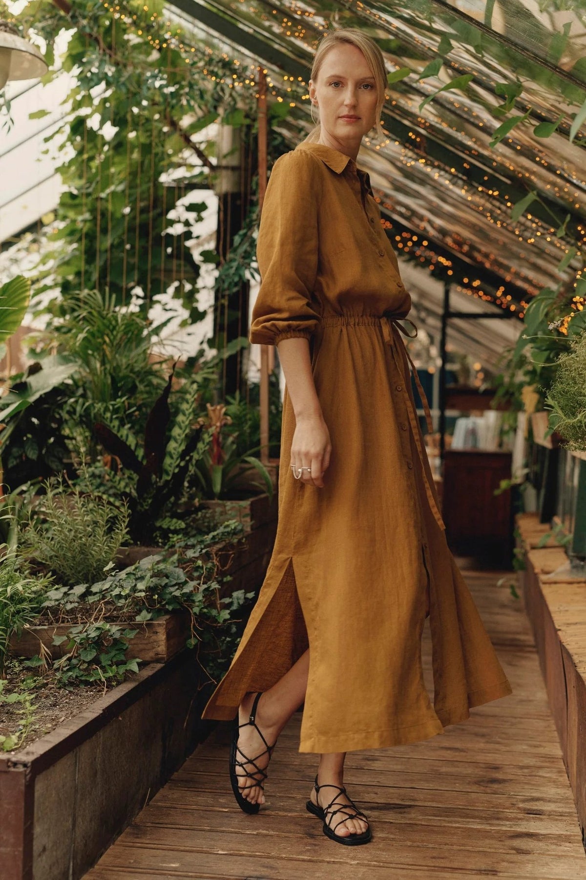 4. Woman in bronze linen Rhapsody dress with button closure and side slits, walking in a greenhouse