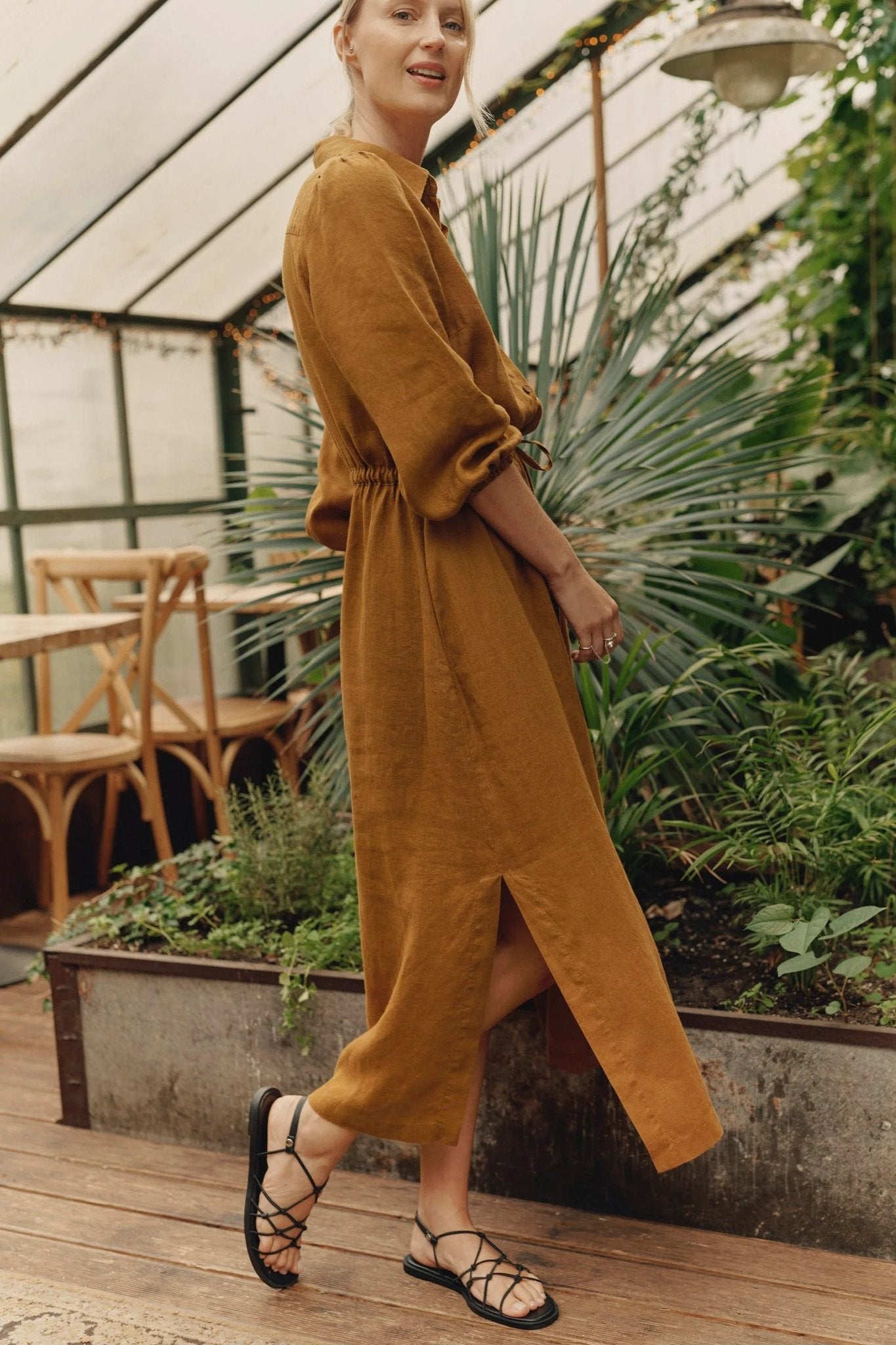 1. Woman wearing bronze linen Rhapsody dress with side slits and adjustable belt, standing in a greenhouse setting