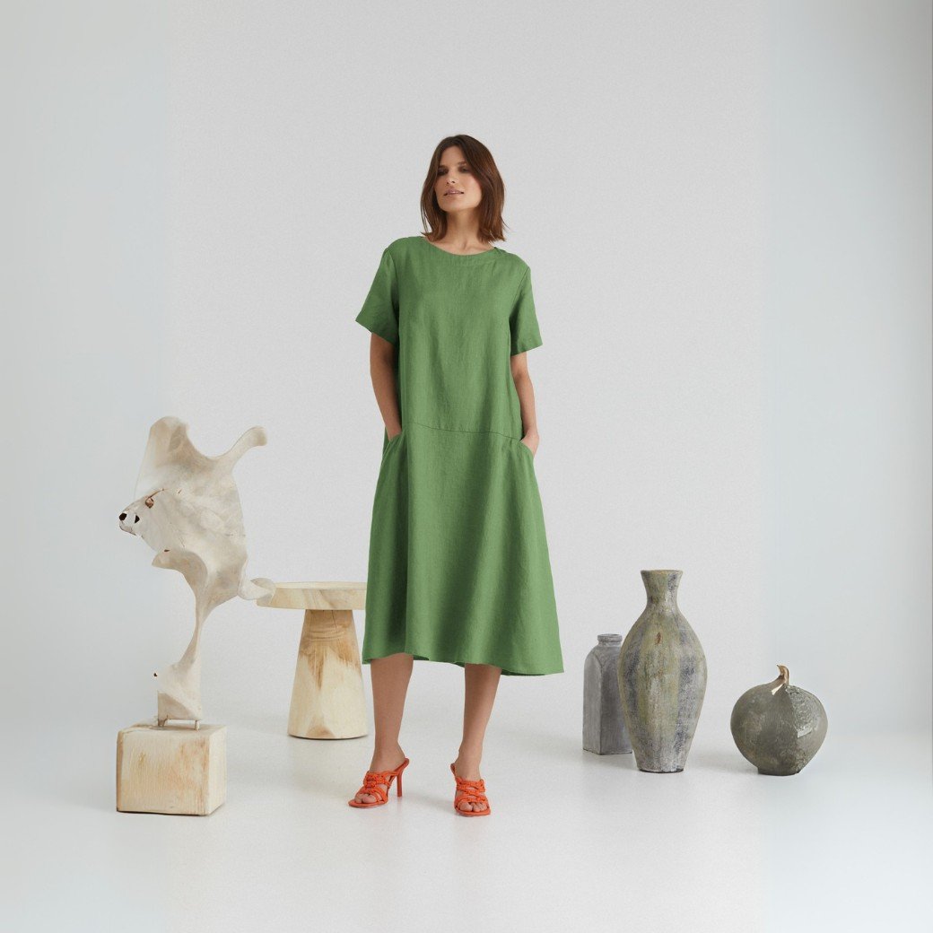 1. Woman wearing green linen A-line dress with short sleeves and round neck, standing in a minimalist setting with decorative vases, showcasing front pockets and relaxed fit