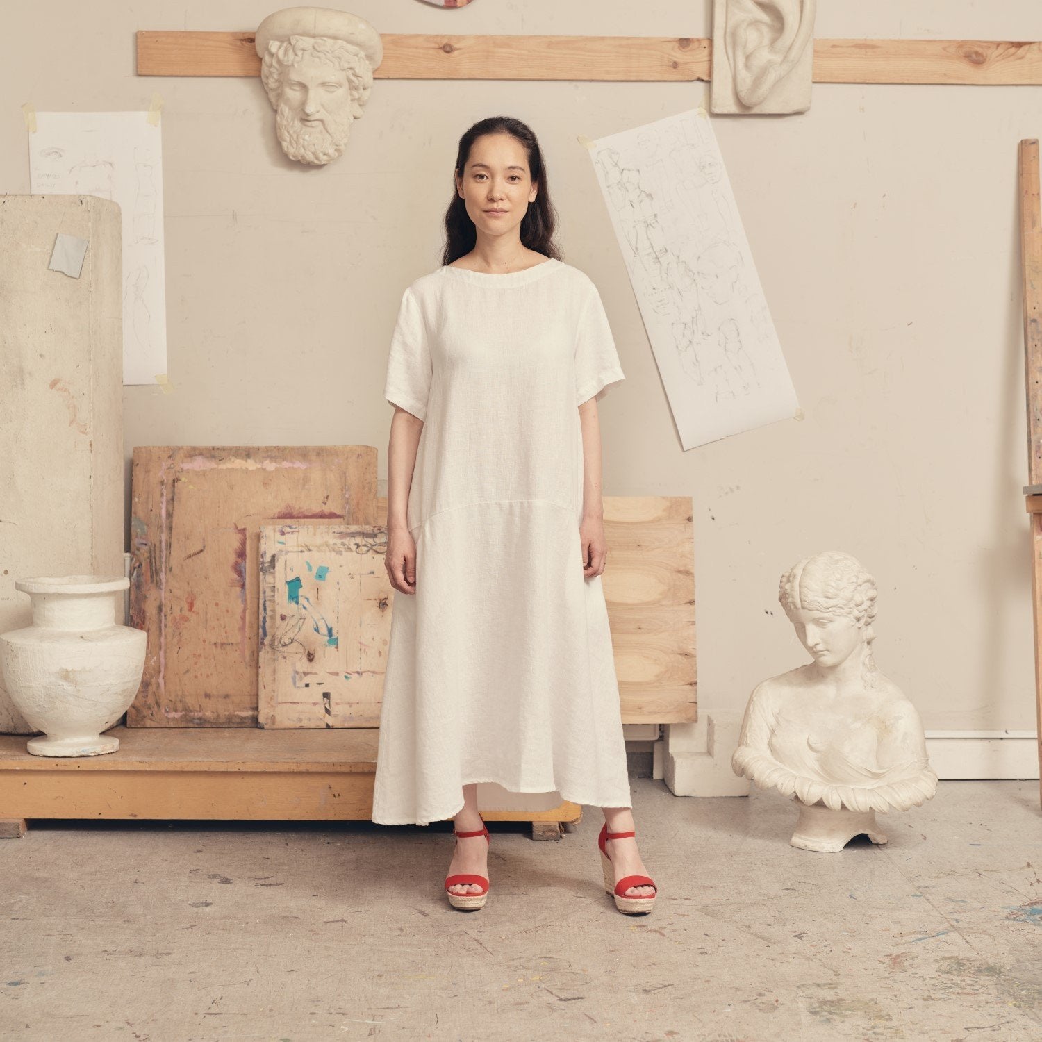 1. Woman wearing optical white linen dress with red sandals in an art studio setting, surrounded by sculptures and sketches