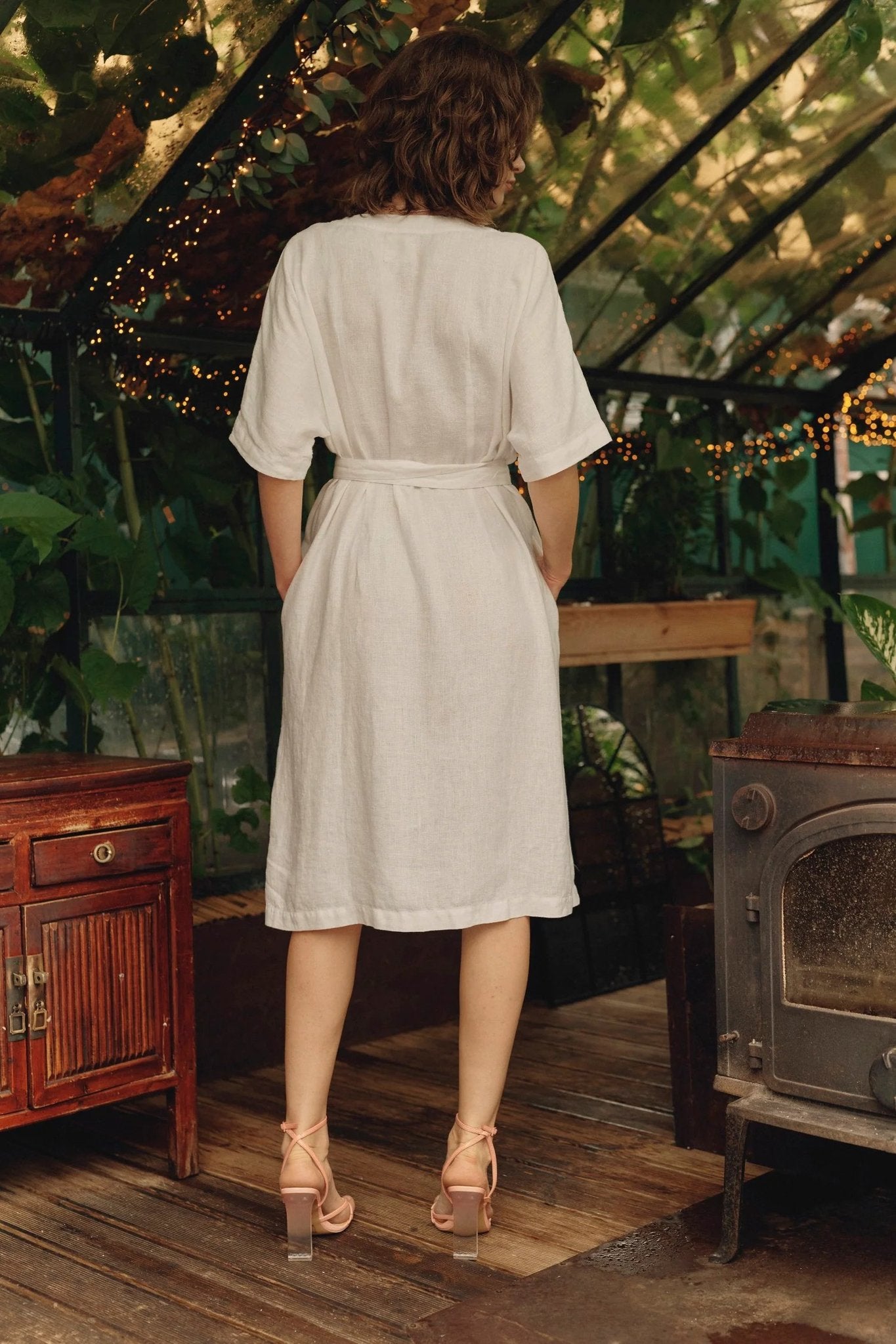 2. Back view of optical white linen wrap dress with tied fastening, worn by woman in greenhouse setting