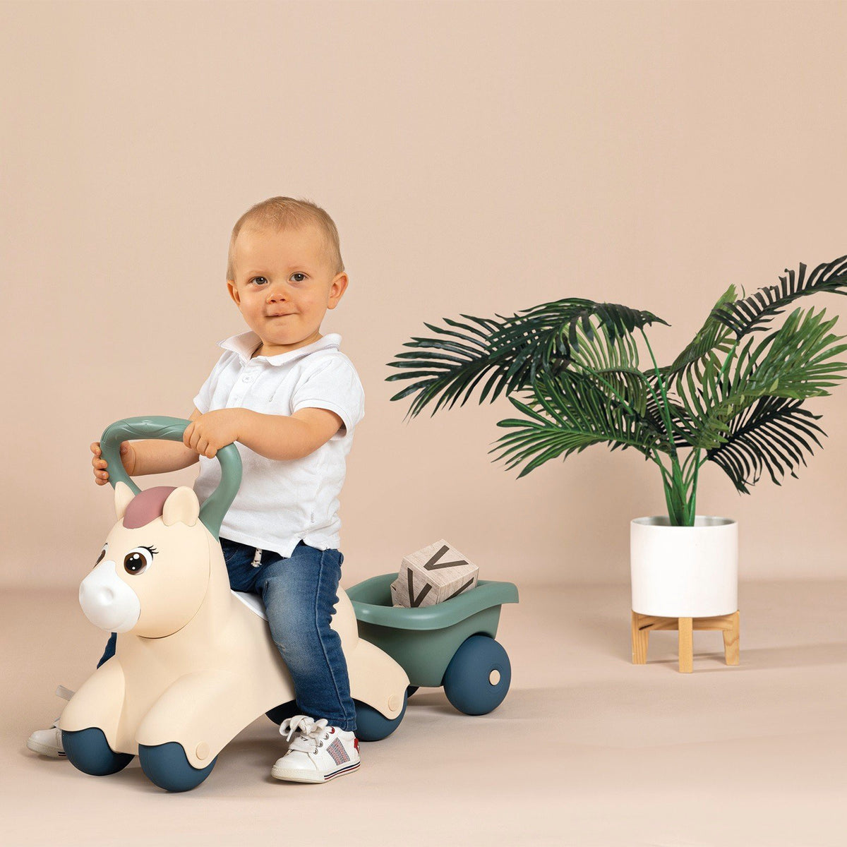 4. Child riding beige pony ride-on with green trailer, smiling in a room with a plant
