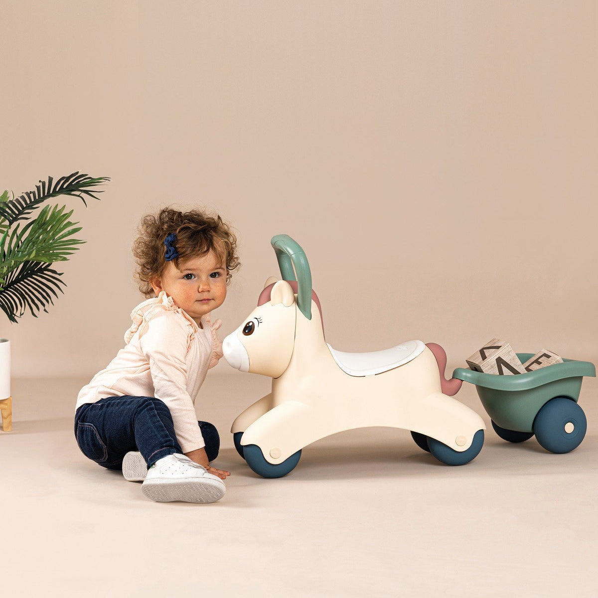 3. Child sitting next to beige pony ride-on with green trailer on a beige background
