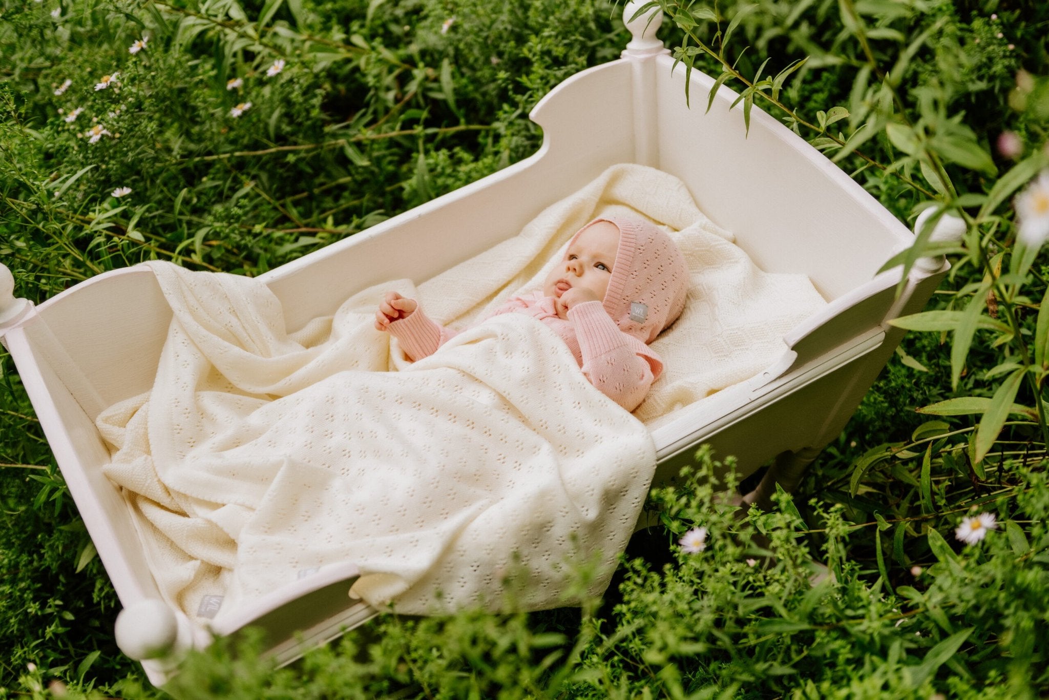 3. Baby in pink merino wool blanket and bonnet lying in a white cradle surrounded by greenery