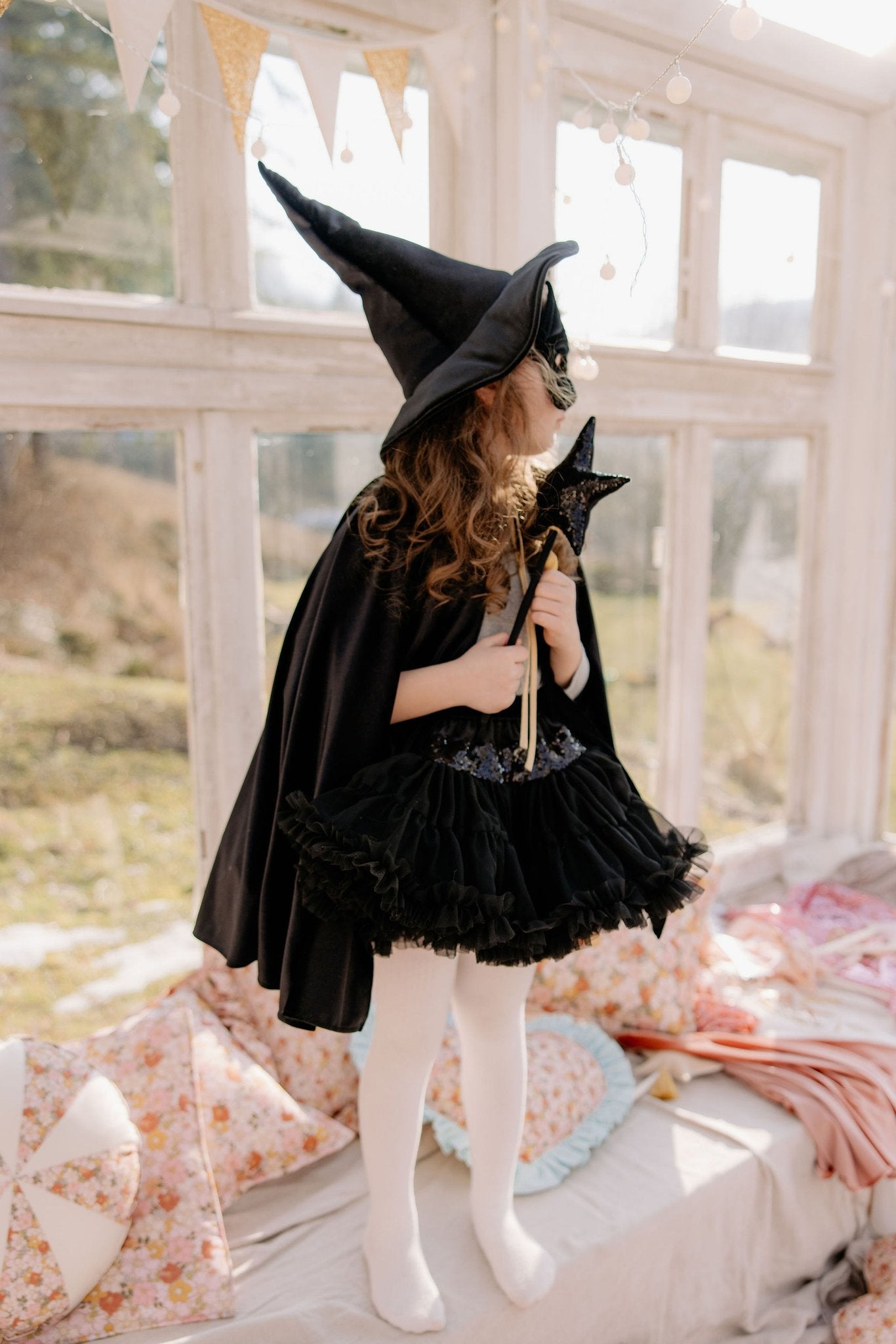1. Child wearing black velour cape and witch hat holding a star wand in a sunlit room