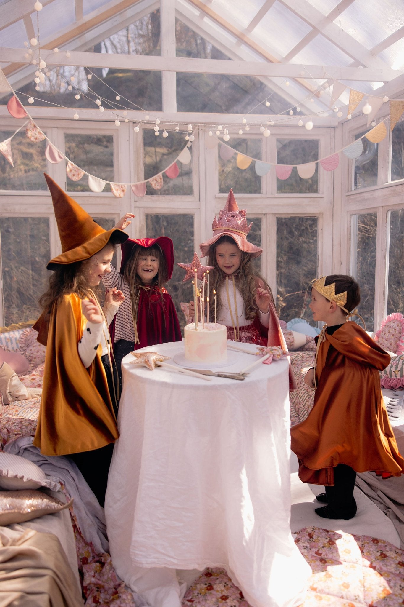 1. Children in capes and hats celebrating around a table with a cake in a bright room