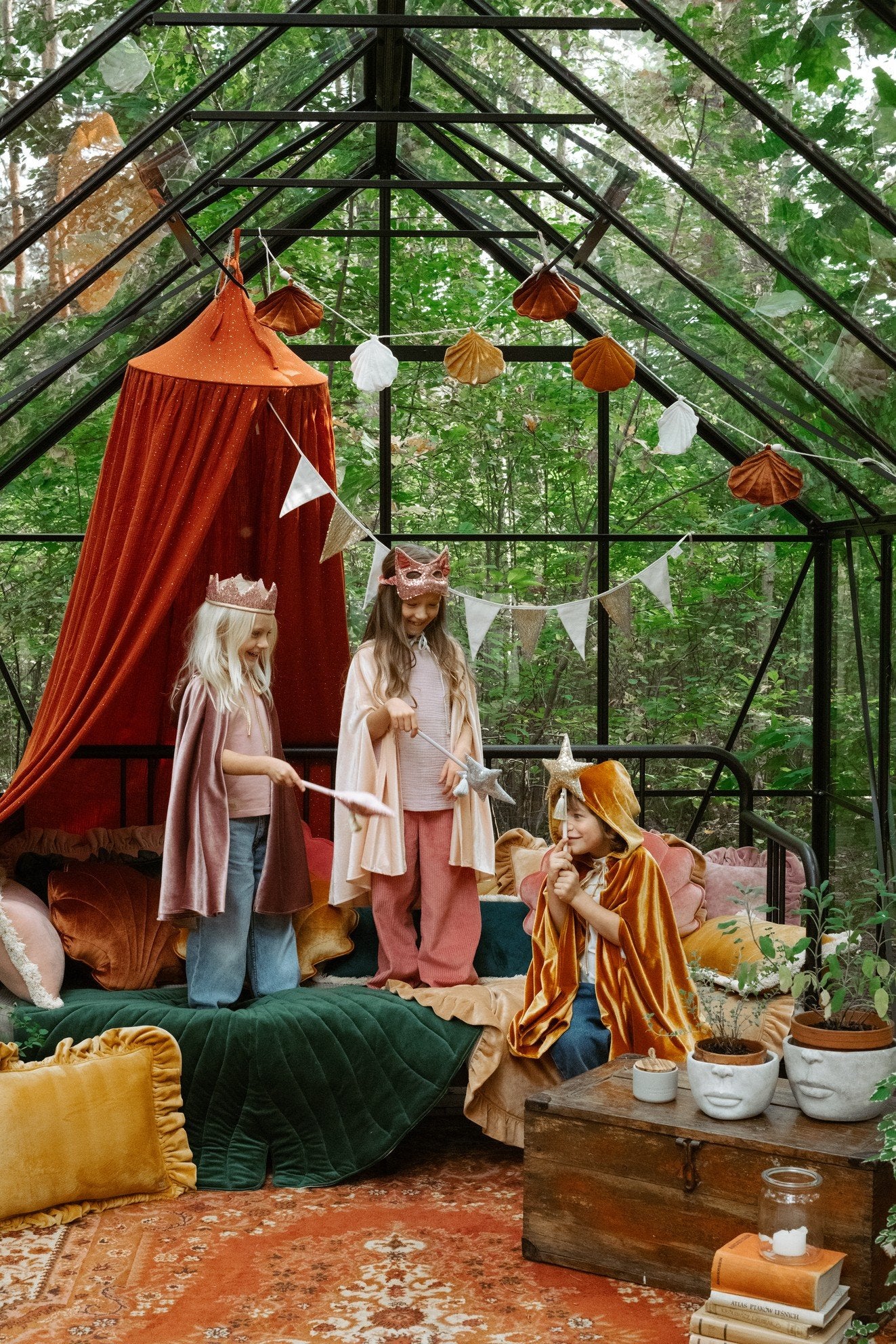 2. Three children in pink and orange capes playing in a decorated greenhouse with banners and cushions