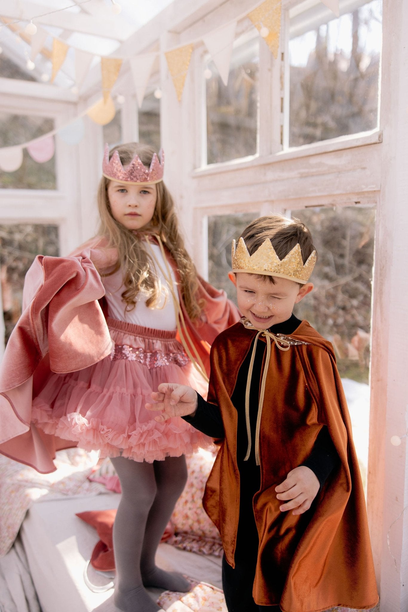 13. Two children in pink and orange capes with crowns in a bright room with bunting