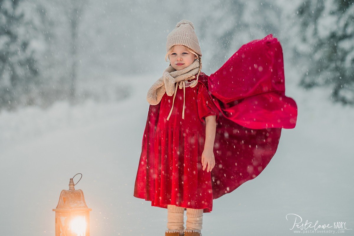 7. Child in red velour cape standing in snowy landscape with lantern, wearing a knit hat and scarf