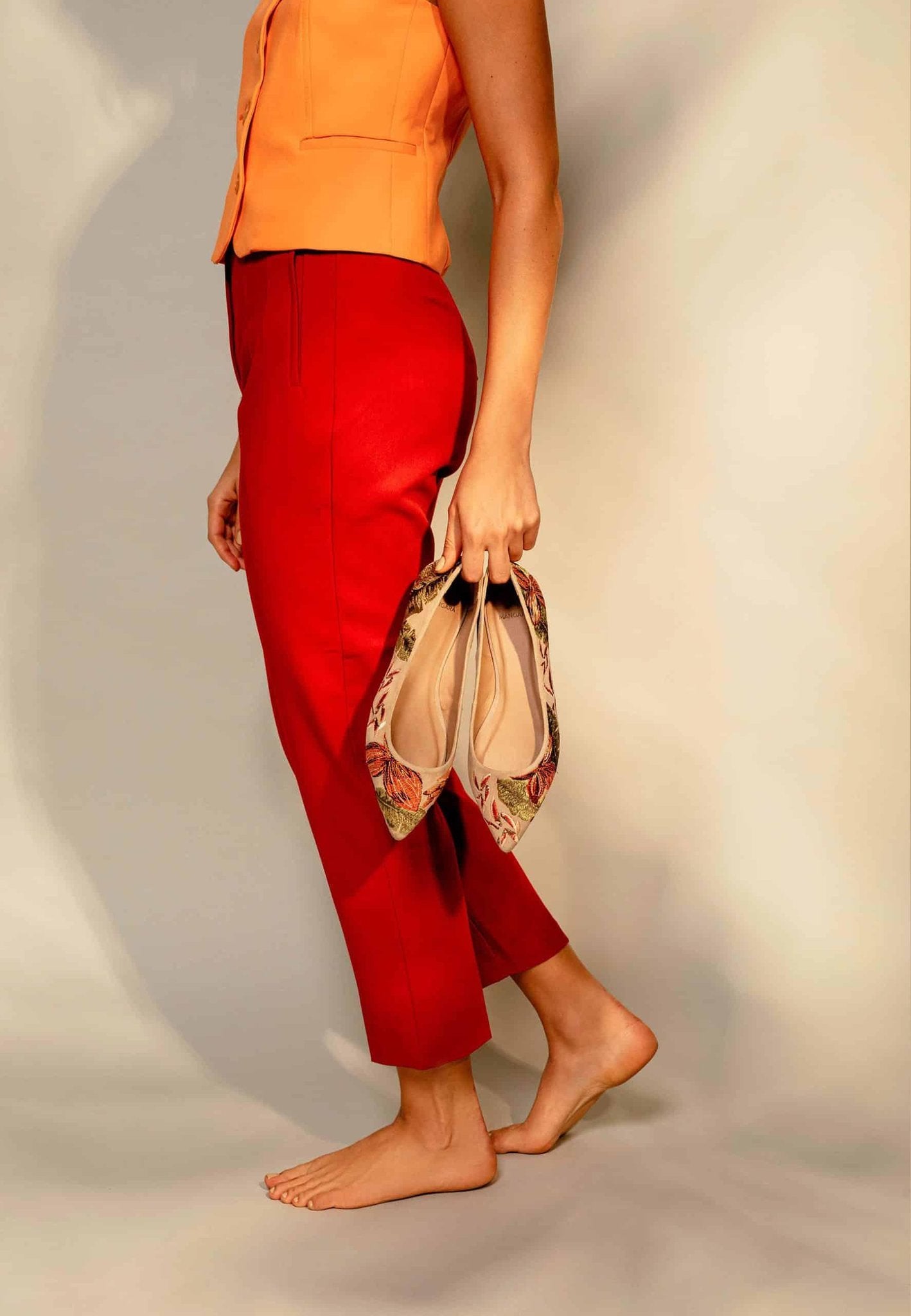 4. Woman holding Mangará Pindaiva Ballerinas in beige suede with floral embroidery, styled with red pants and orange top