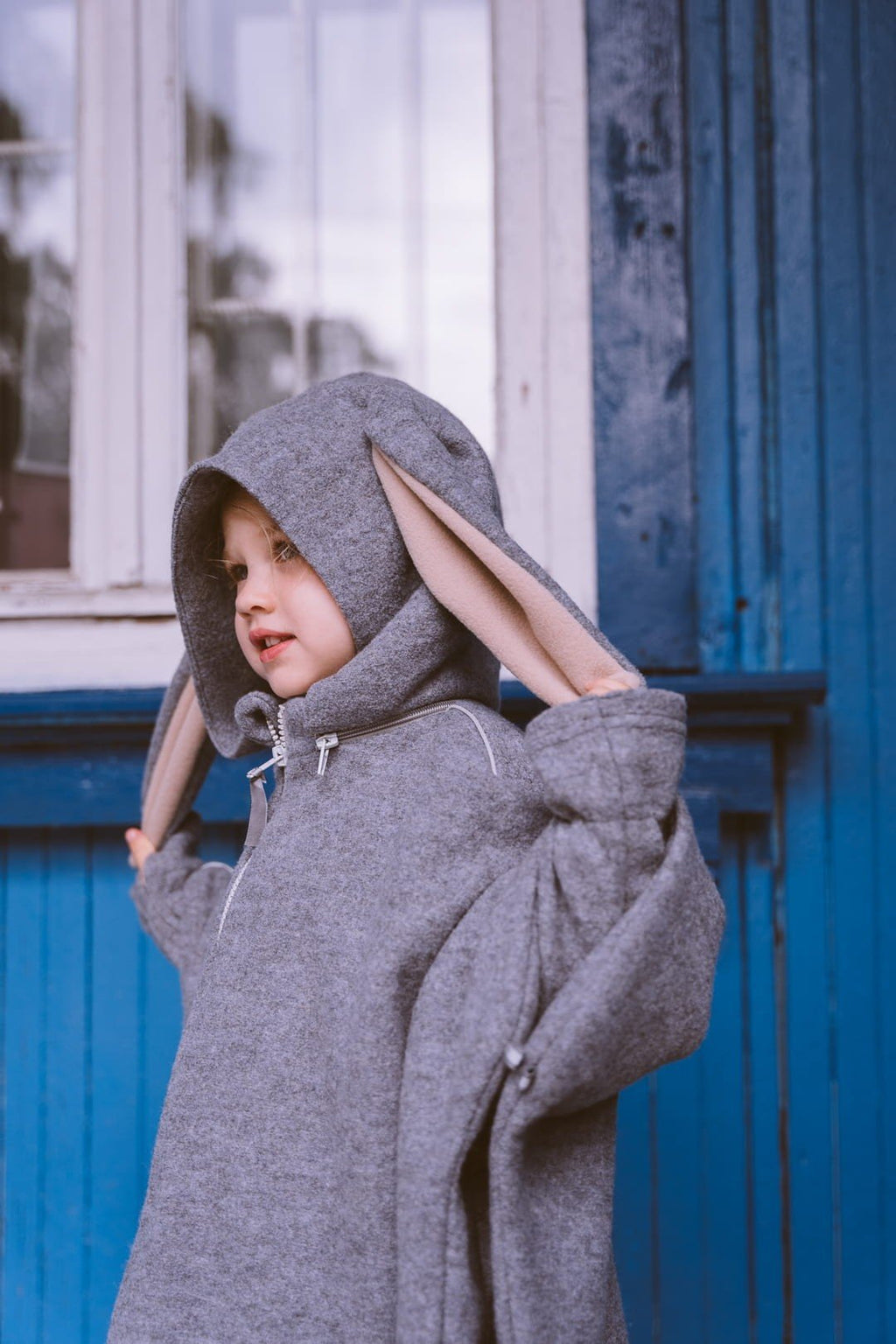 4. Child wearing grey merino wool poncho with bunny ears standing by a blue house