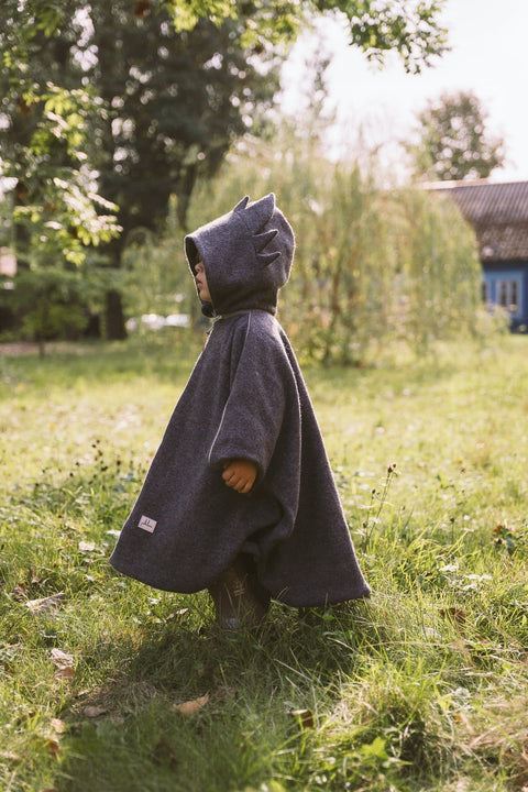 4. Child walking in grassy field wearing grey merino wool poncho with dragon hood, side view