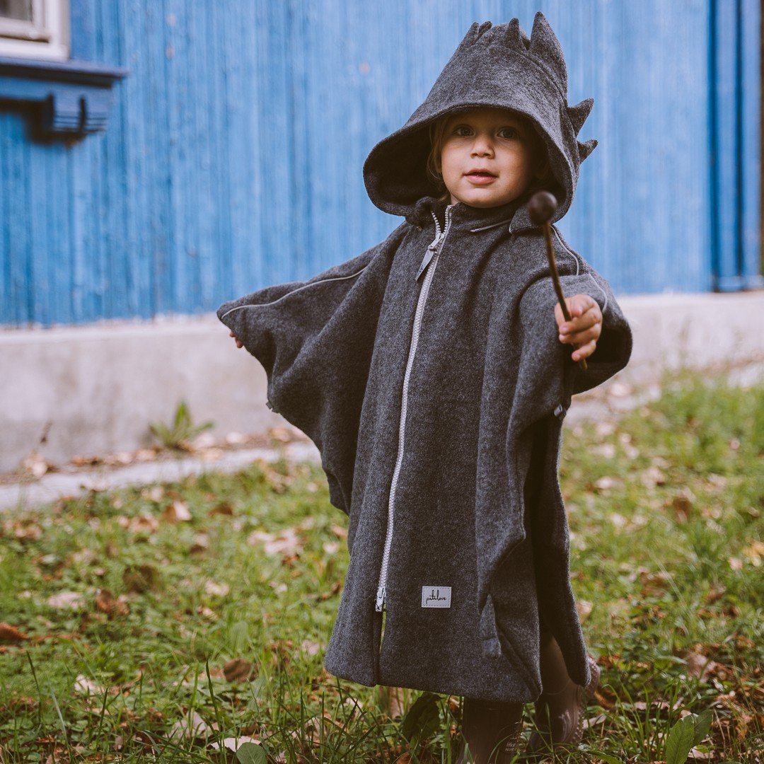 1. Child wearing grey merino wool poncho with dragon hood outdoors, holding a stick, in front of a blue building