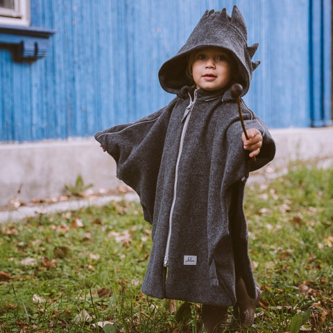 1. Child wearing grey merino wool poncho with dragon hood outdoors, holding a stick, in front of a blue building
