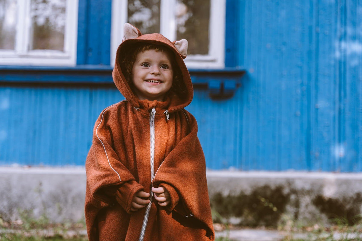 2. Smiling child in brown merino wool poncho with fox ears hood in front of blue house