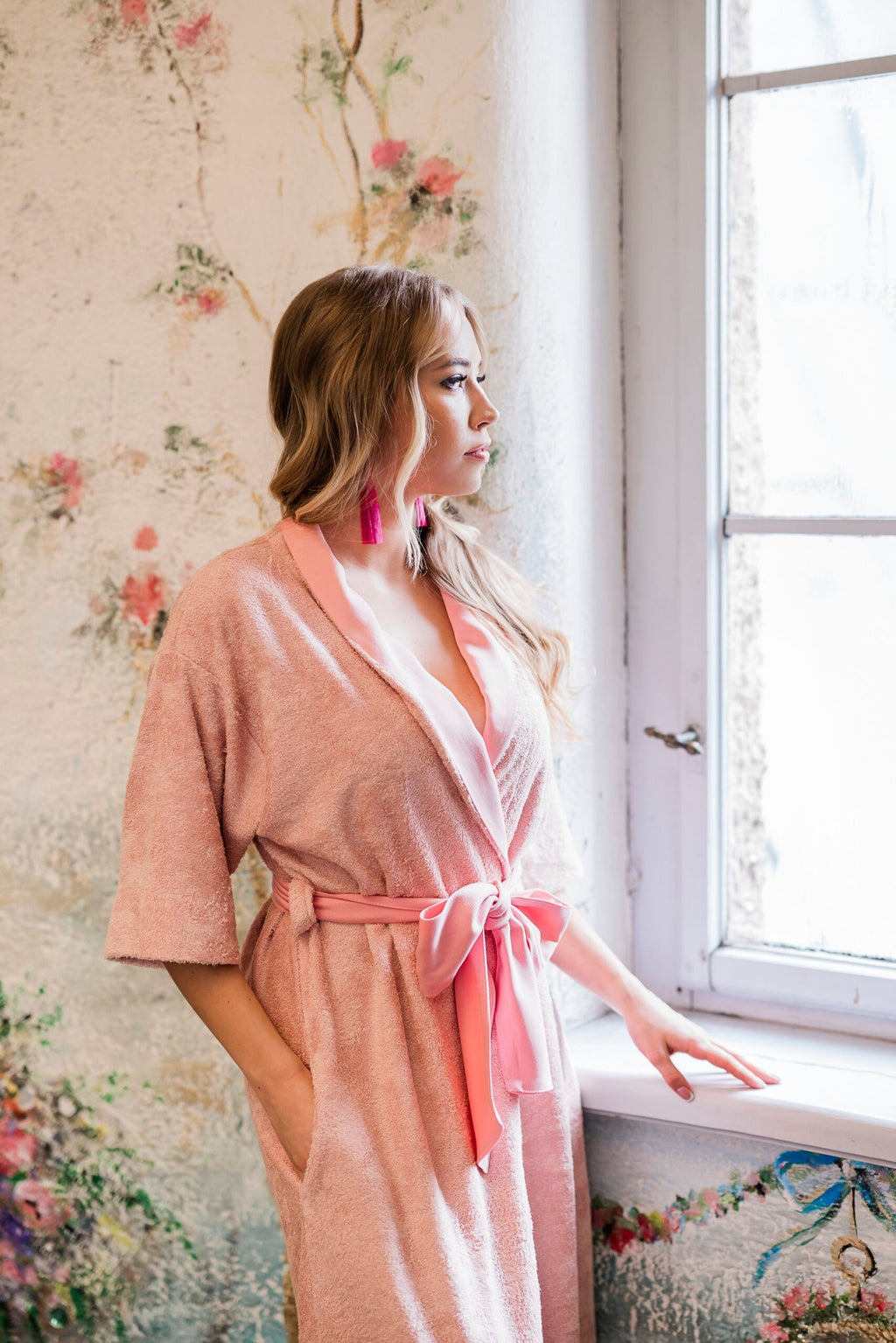 1. Woman in pink bamboo terry robe with shawl collar and bow belt looking out window in floral room