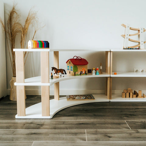 2. Corner view of modular Montessori shelf with three white shelves and natural wood supports, decorated with toys and books, on wooden flooring.