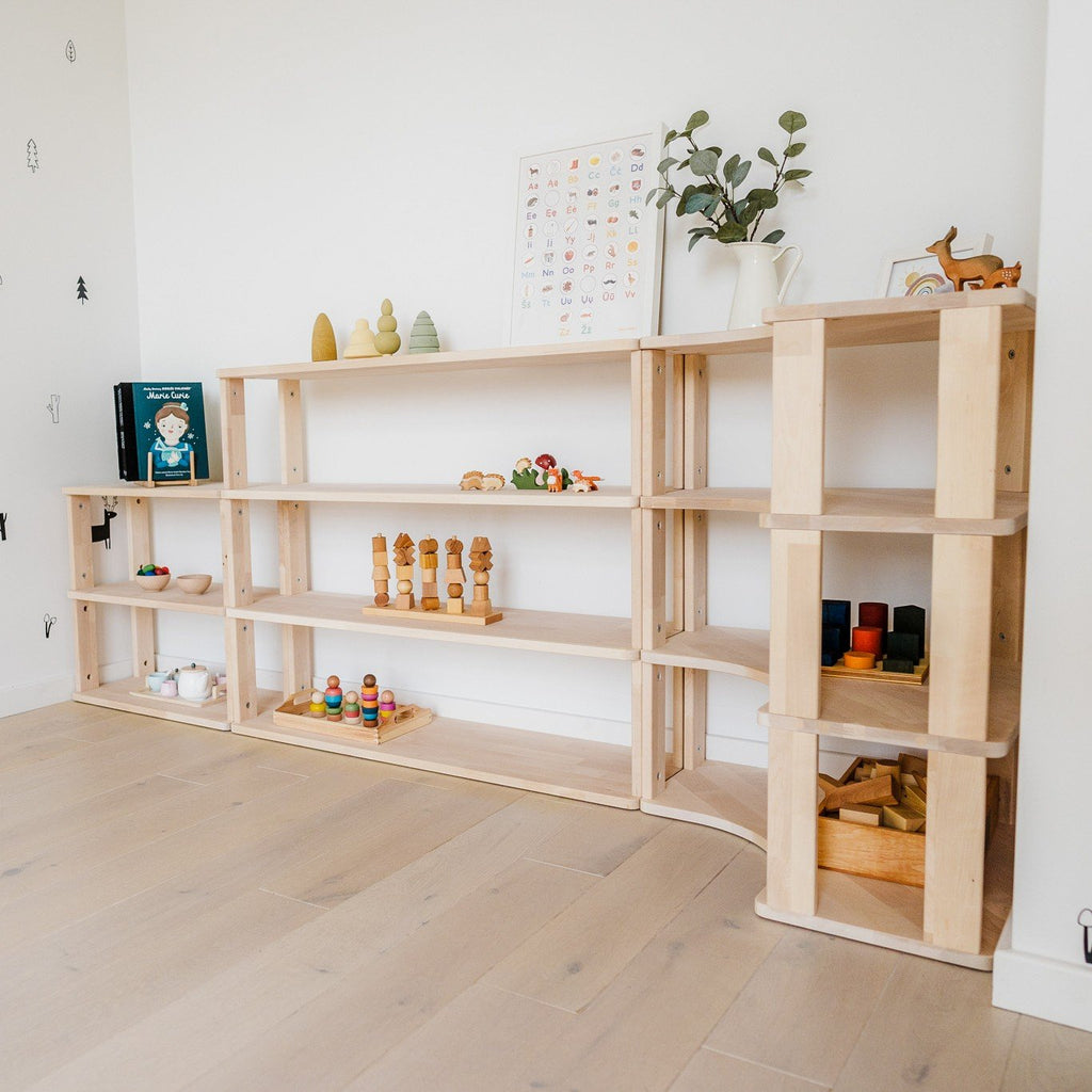 6. Modular Montessori shelf with natural wood finish, displaying toys and books, set against a white wall on light wooden flooring.